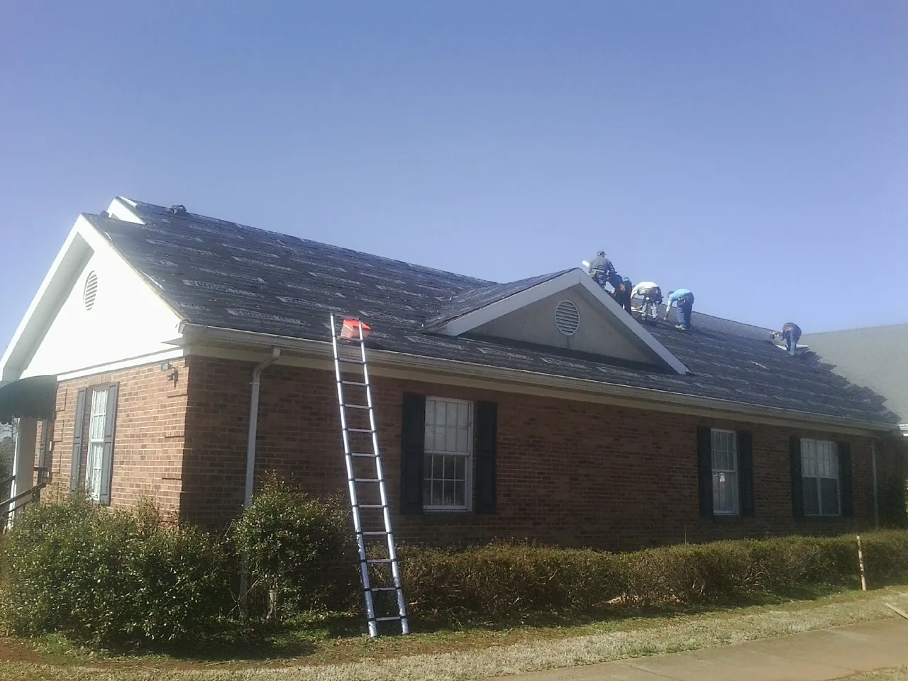 Crew tearing shingles off a brick ranch roof with a ladder set — Metal Roof Repair work by our crew in Cincinnatus
