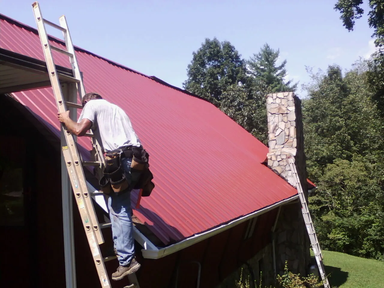 Roofer on a ladder beside a red metal roof and stone chimney — Soffit & Fascia Repair by Roofing Brothers in Cincinnatus
